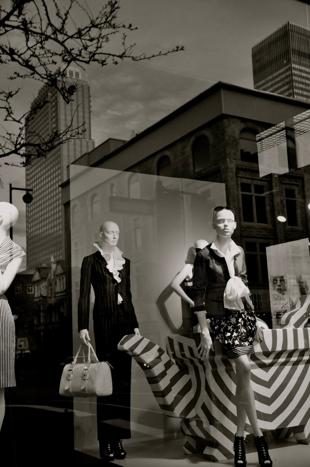 Black and white photo of clothing store window display with mannequins and bold striped backdrop in urban setting.