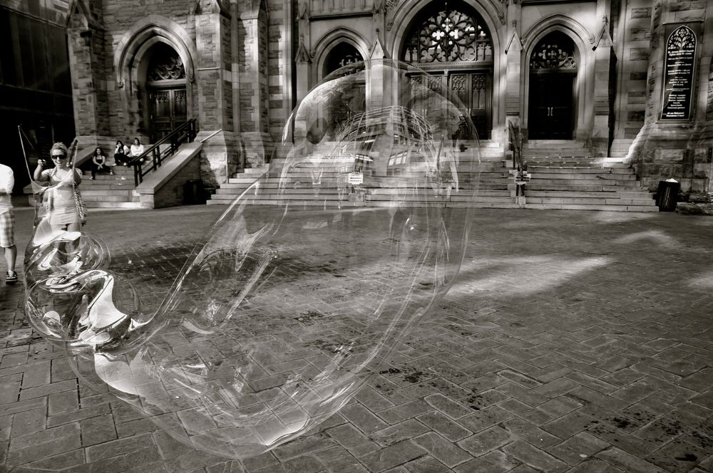 Ethereal soap bubble floating near stone archways in an urban square, captured in monochrome.