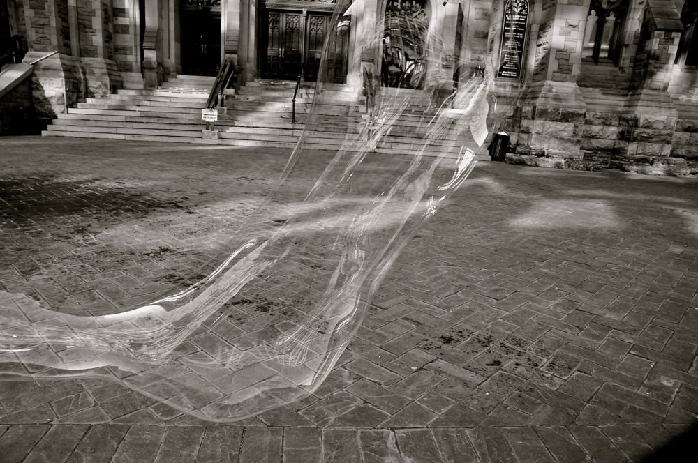 Ghostly trail of a soap bubble's path photographed against historic architecture in black and white.