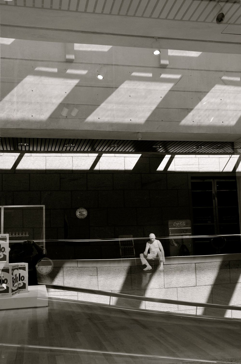 A person sits alone in a sunlit gymnasium with dramatic shadows cast across the floor from overhead skylights.