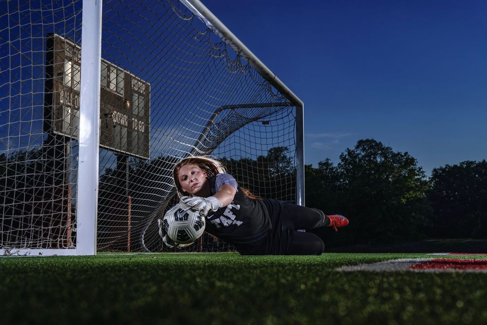 Soccer goalkeeper makes a diving save on artificial turf field with goal post in frame.