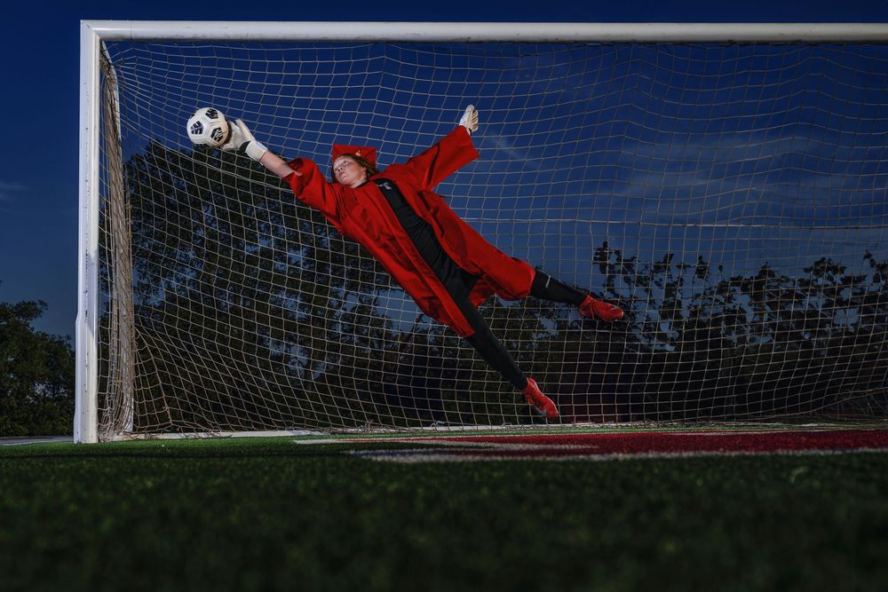 Soccer goalkeeper in red uniform making a dramatic diving save attempt against a blue net at sunset.