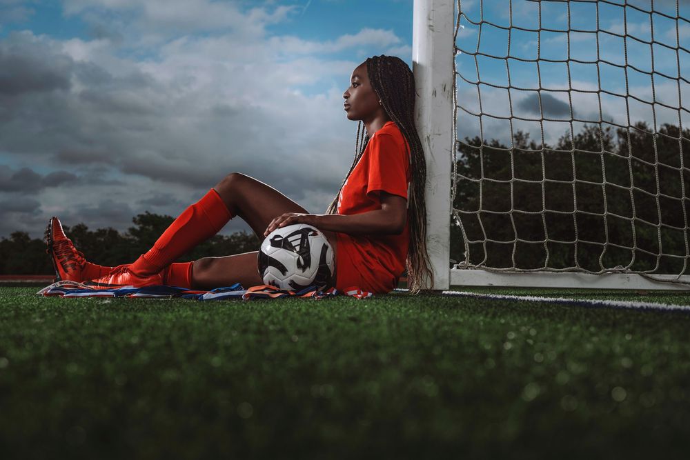 Soccer player in orange uniform rests against goal post with ball on grass field during cloudy day.