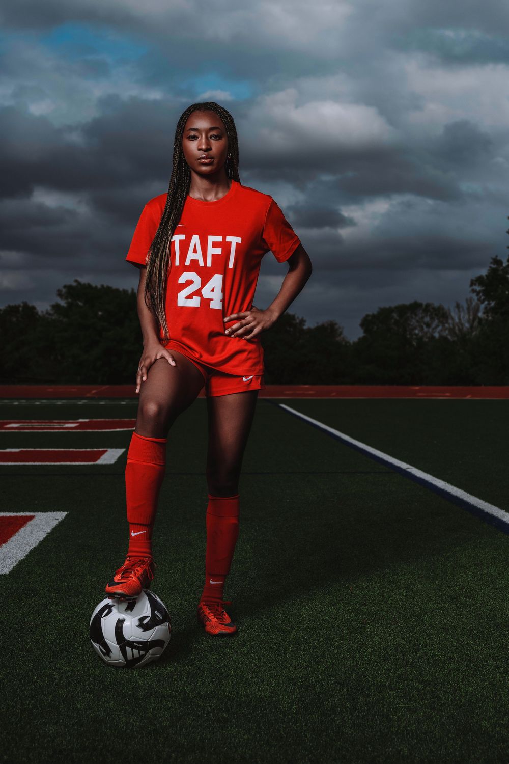 Athlete in red Taft jersey number 24 poses with soccer ball on turf field under dramatic stormy sky at dusk.