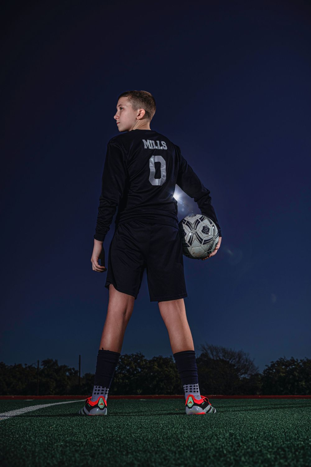 Youth soccer player in black uniform holding ball stands on turf field under dark evening sky.