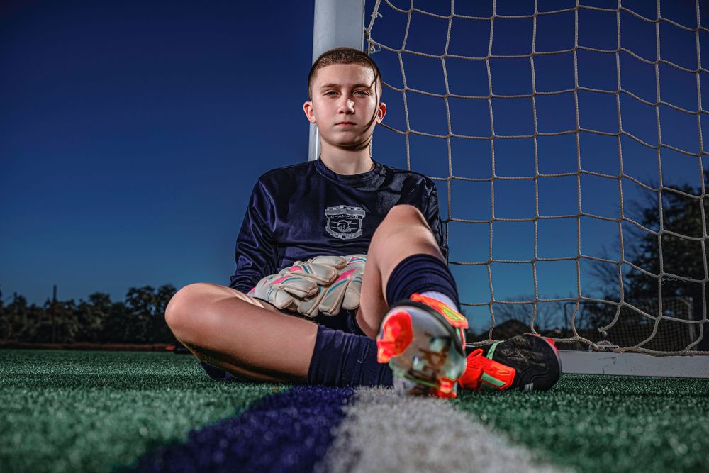 Soccer player sits on artificial turf field in front of goal net wearing dark uniform and orange cleats at dusk.
