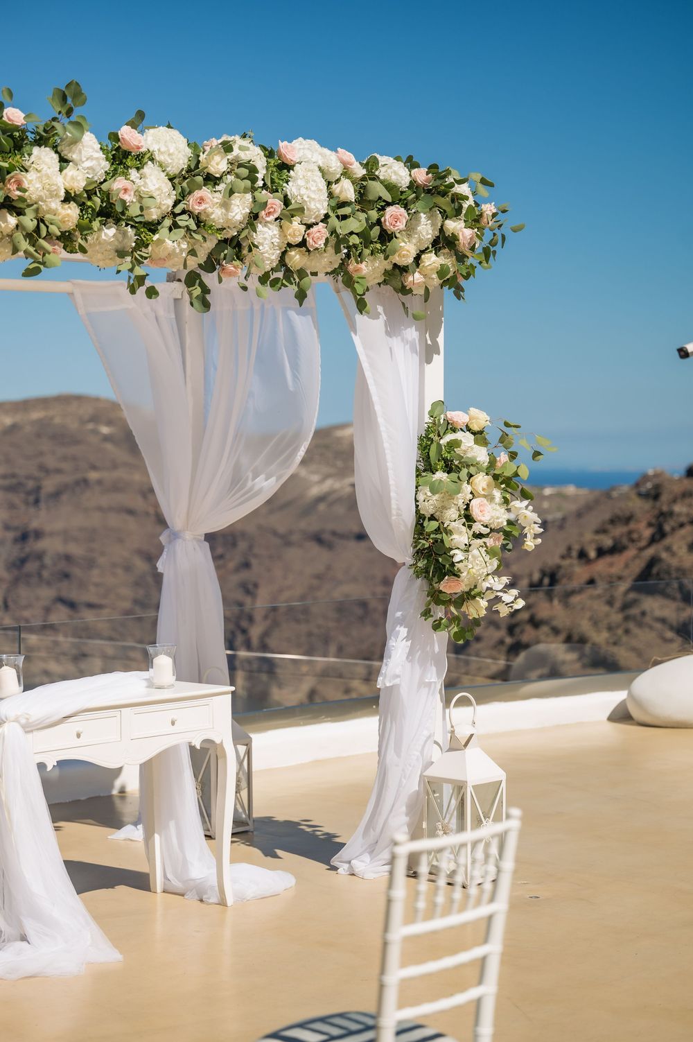 Elegant white wedding arch with floral arrangements overlooking mountainous landscape at outdoor venue in Santorini.