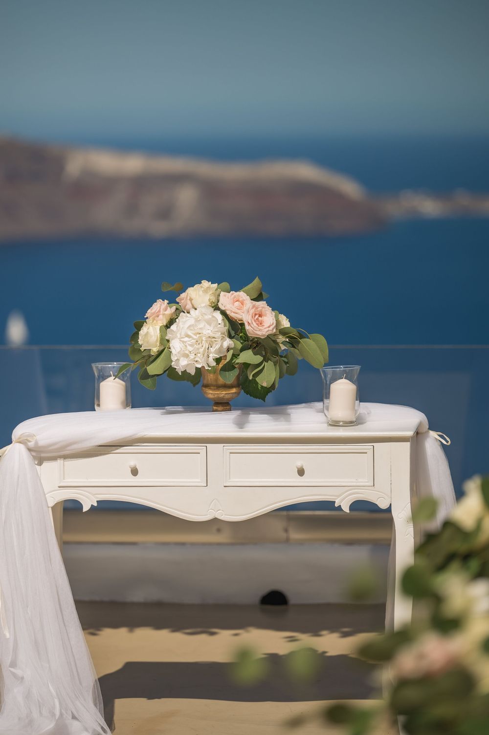Elegant white console table with floral centerpiece and candles overlooking Mediterranean sea views in Santorini.