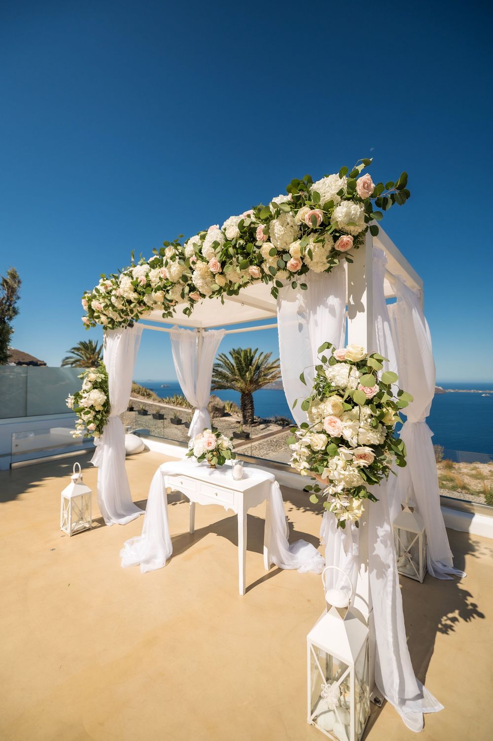 White draped wedding arbor decorated with roses and greenery against stunning ocean backdrop.