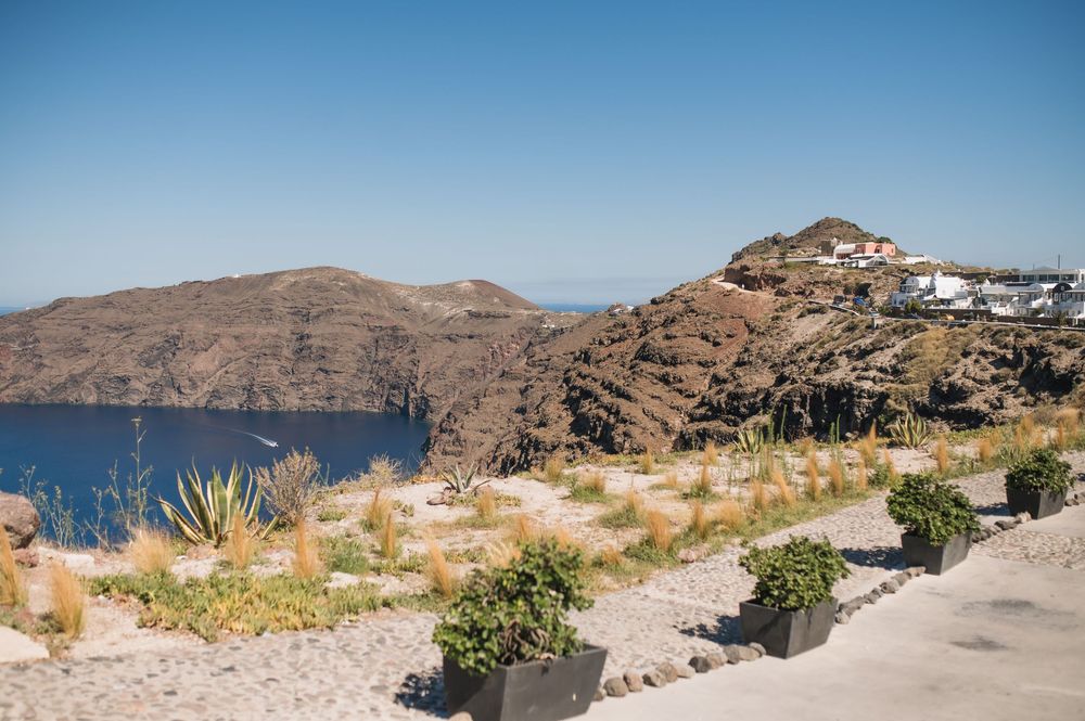 A scenic view of rocky cliffs and Mediterranean coastline in Santorini with blue waters and coastal vegetation in foreground.