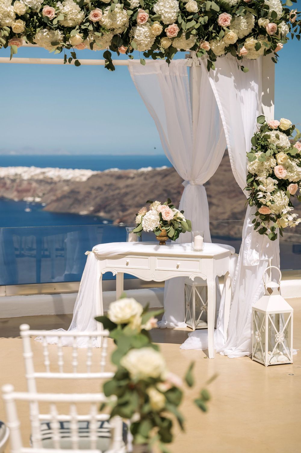 Wedding ceremony setup with white floral arch, lanterns, and chairs against a stunning ocean backdrop.