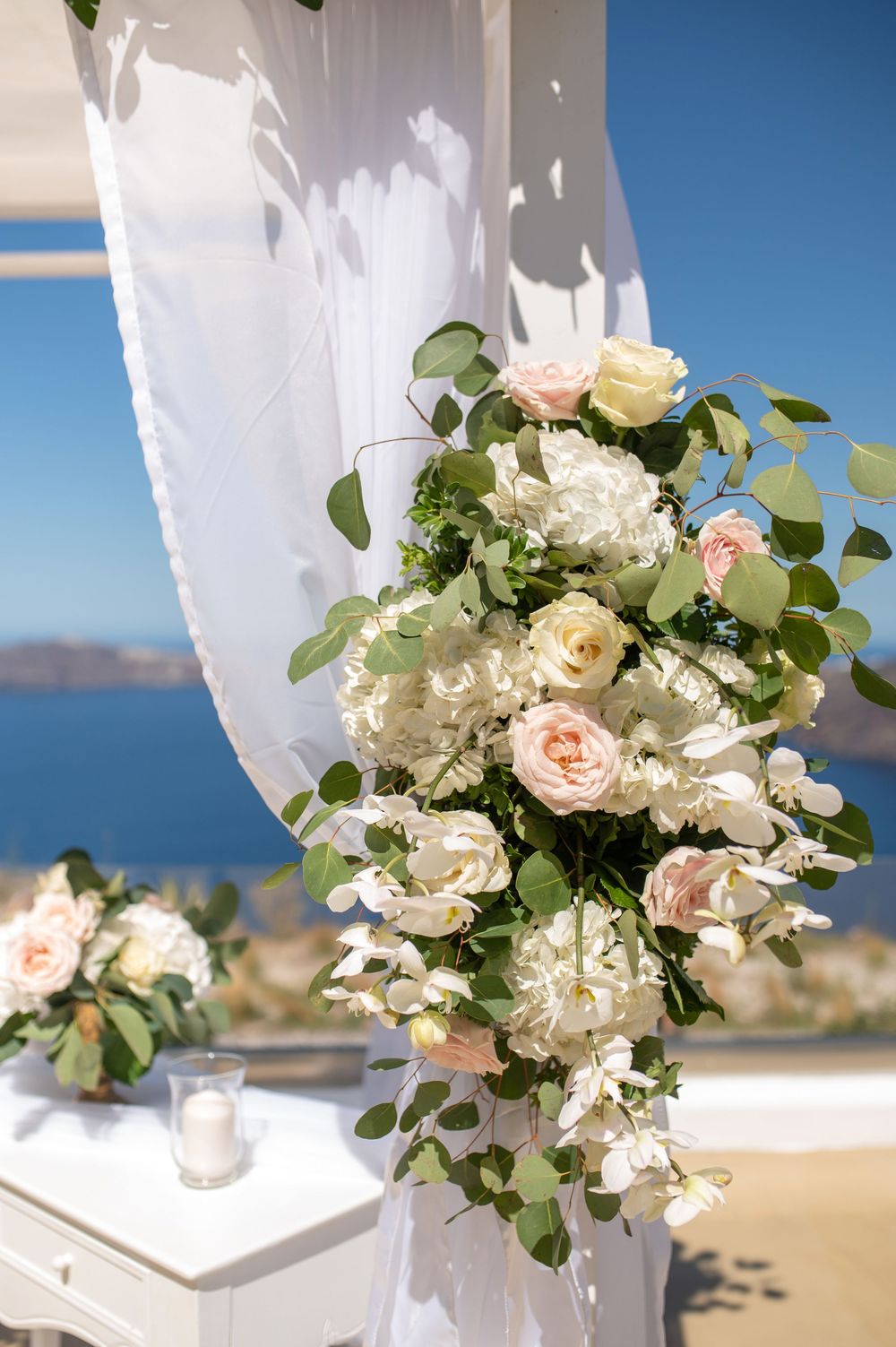Elegant floral arrangement with white hydrangeas and pale pink roses adorning a white curtain against a scenic ocean backdrop.