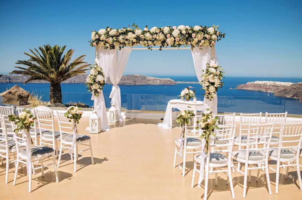 Wedding ceremony setup on a terrace overlooking the Mediterranean Sea with white chairs and floral arch in Santorini, Greece.