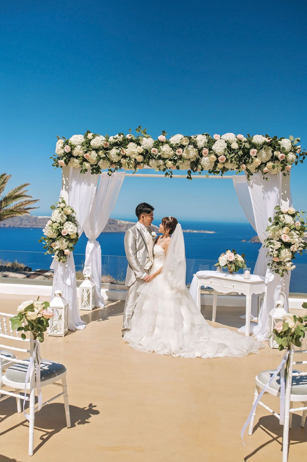 Beautiful seaside wedding ceremony setup with white floral arch, sheer drapes and ocean views in Mediterranean location.
