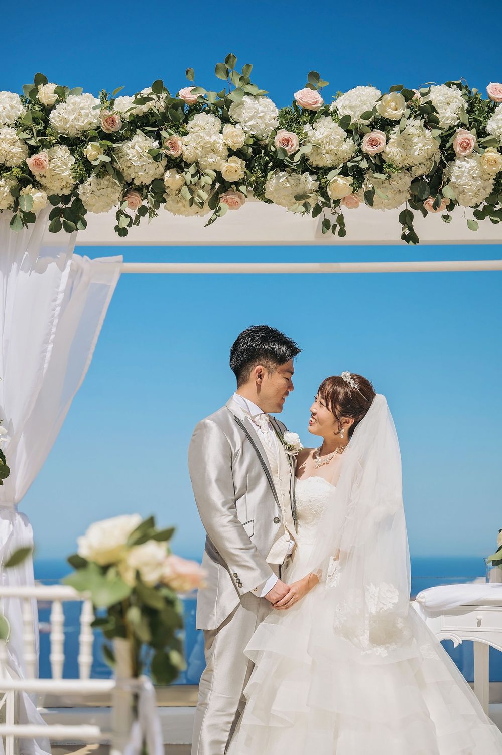 Beautiful garden wedding arch with white roses and flowing drapes against blue sky over oceanfront ceremony space.