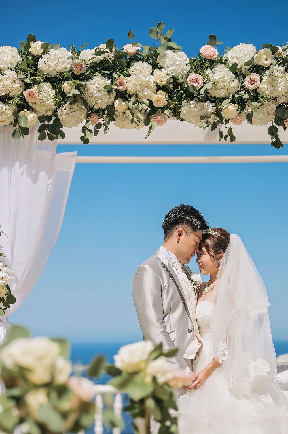 Romantic outdoor wedding ceremony setup with white floral arch and sheer drapes against blue ocean backdrop.