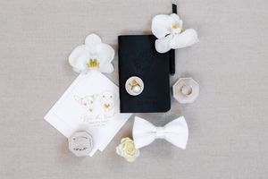 Minimalist wedding flatlay with white flowers and black accents on grey background.
