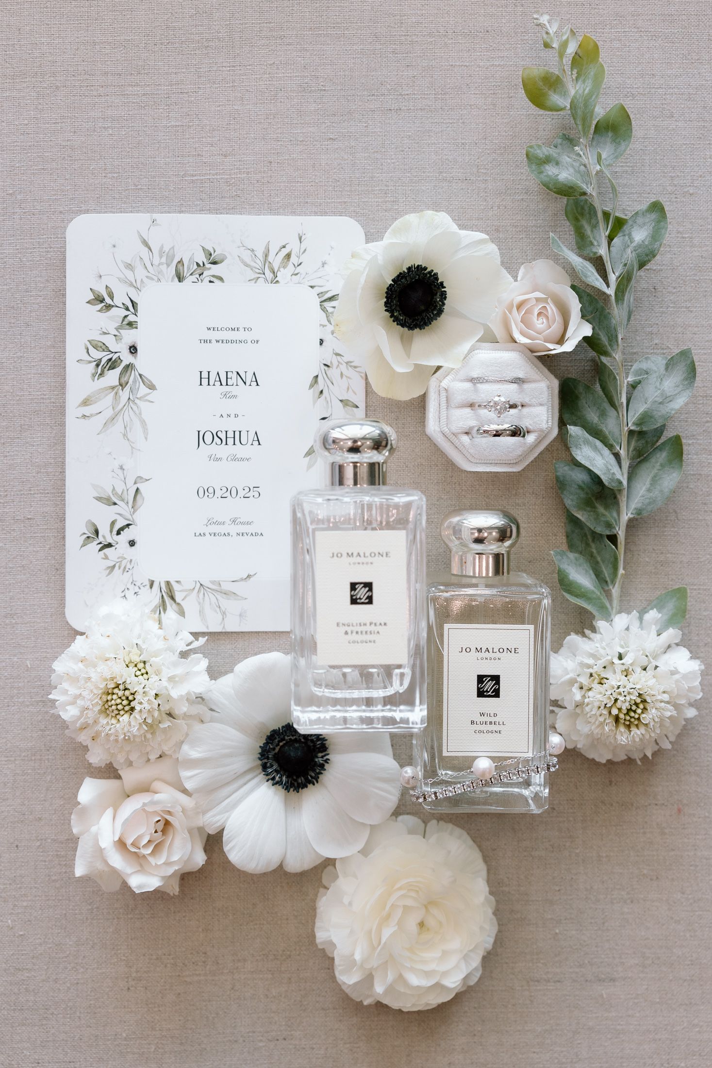 Elegant wedding flatlay with white flowers, perfume bottles, and invitation cards arranged on neutral linen background.
