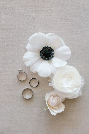 Three silver wedding rings arranged with delicate white flowers including an anemone and roses on light linen background.