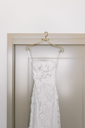 White lace wedding dress hanging on a gold hanger against a neutral wall.