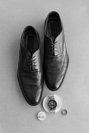 Black leather dress shoes and a silver watch arranged on a light gray background in a minimalist style.