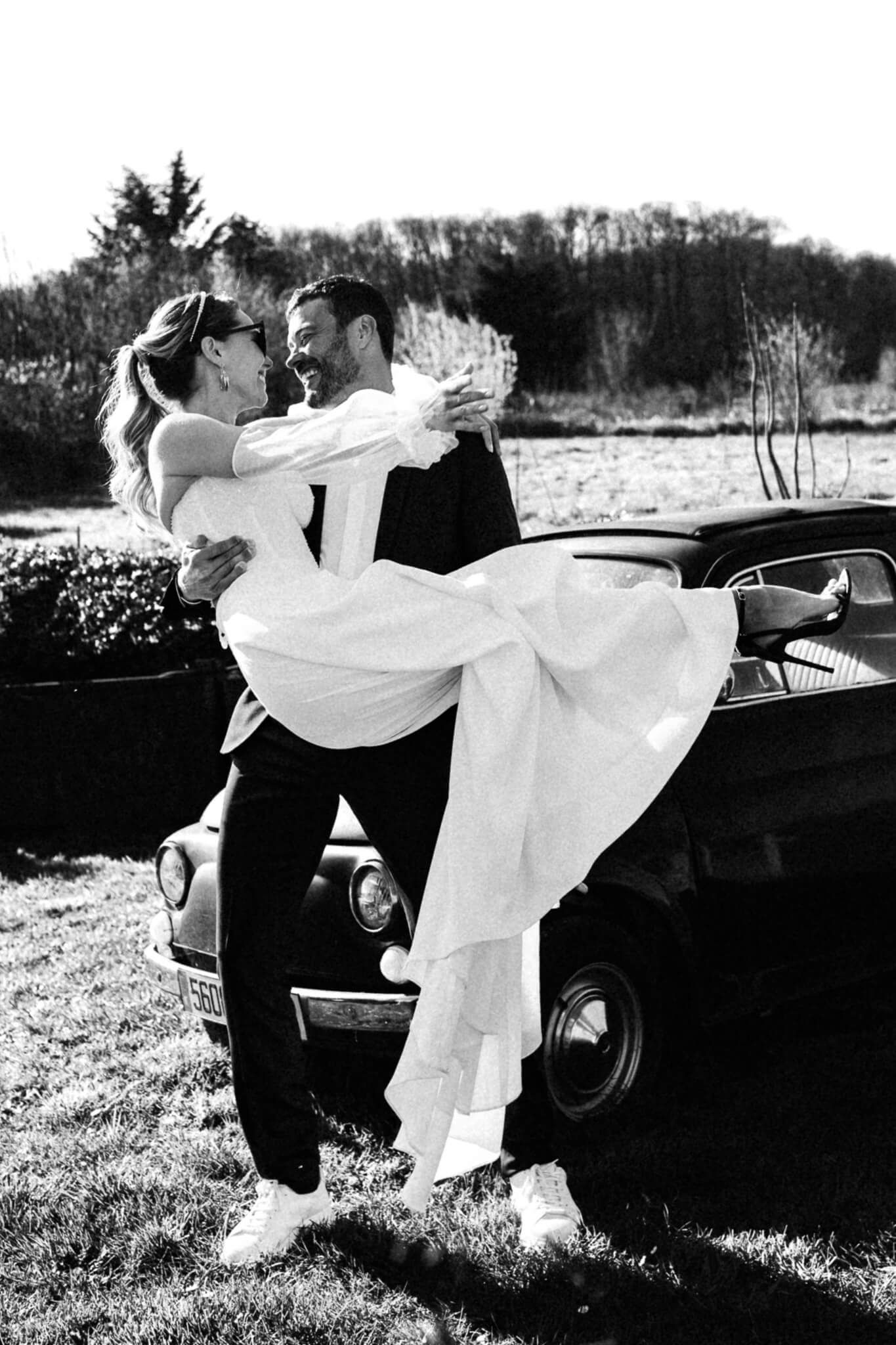 Mariés en noir et blanc rétro devant une voiture ancienne