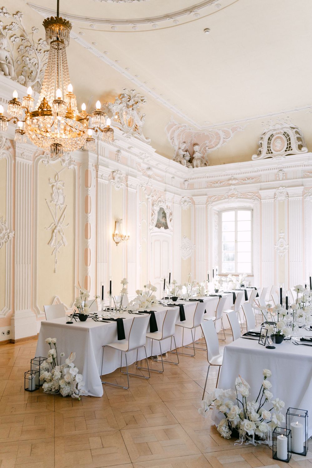 Elegant white ballroom with crystal chandelier and minimalist table settings for wedding reception in baroque palace interior.