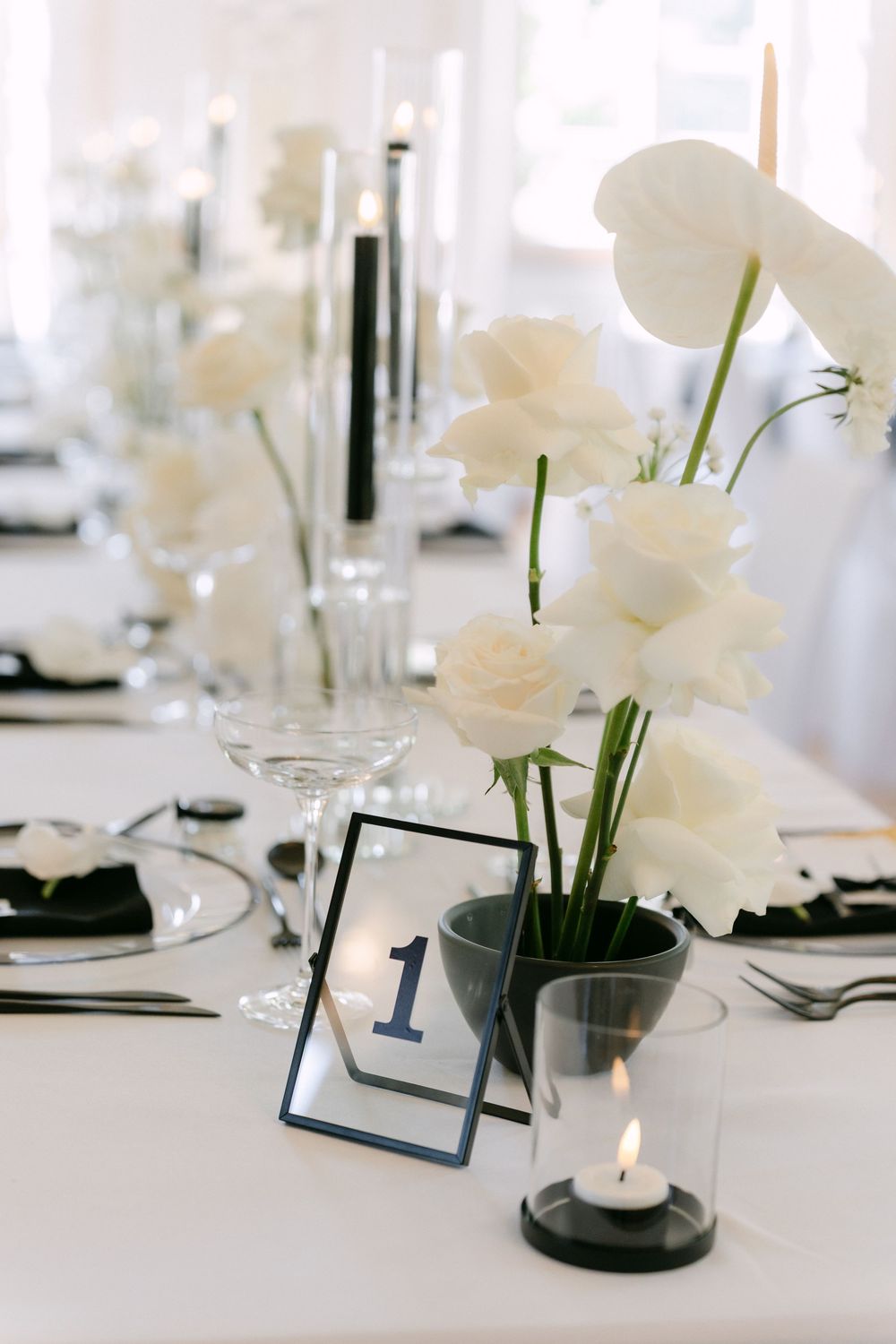 Elegant wedding table setting with white roses in vase, table number one, and glowing candle on crisp white tablecloth.