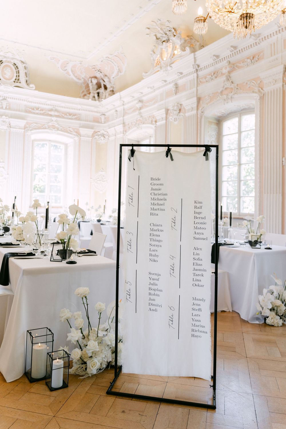 Elegant wedding seating chart display in ornate white room with crystal chandeliers and floral arrangements on round tables.