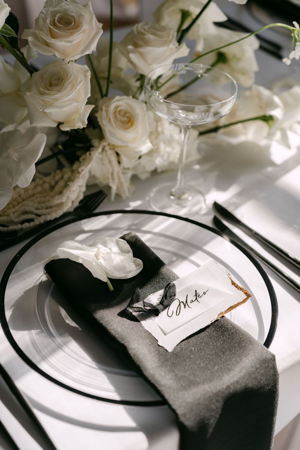 Elegant wedding place setting with white roses, crystal glass, black napkin and name card on pristine white tablecloth.