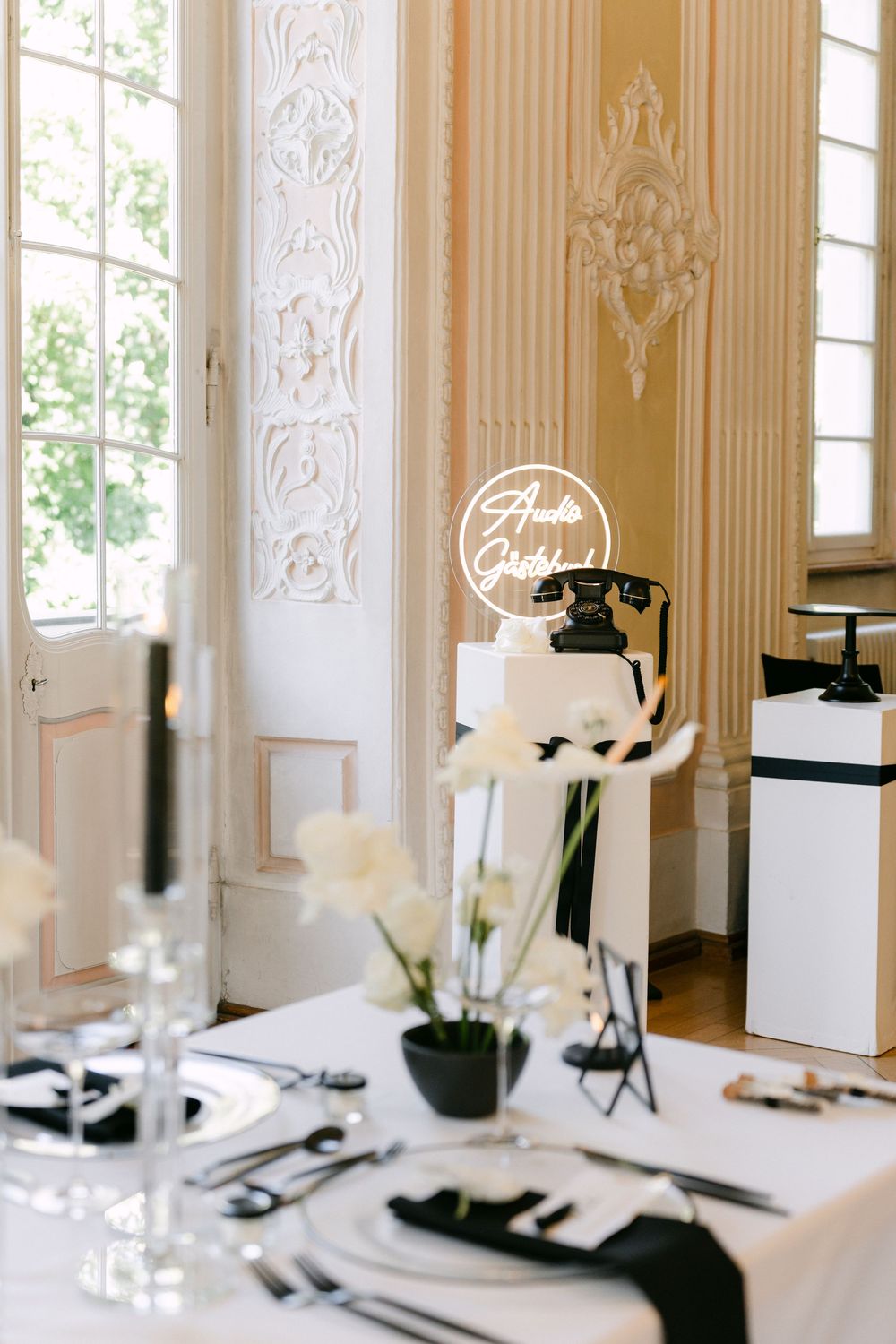 Elegant table setting with vintage black rotary phone and white floral decor in a room with ornate wall panels and windows.