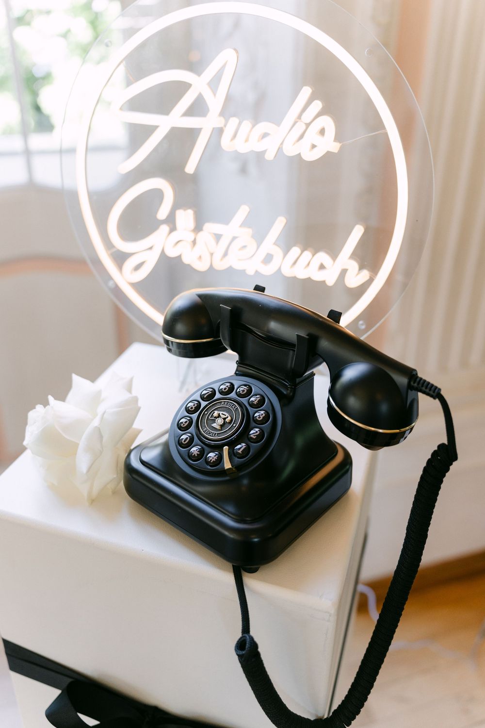 Vintage black rotary telephone displayed with illuminated 'Audio Gästebuch' neon sign on white surface.