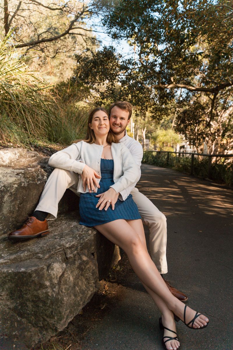 man sitting on a rock and hugging his girlfriend who is sitting in front of him and both are looking at the camera during a photoshoot