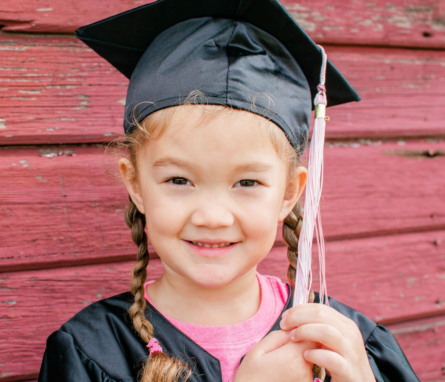 Young graduate in black cap and gown with braided hair against red wall.
