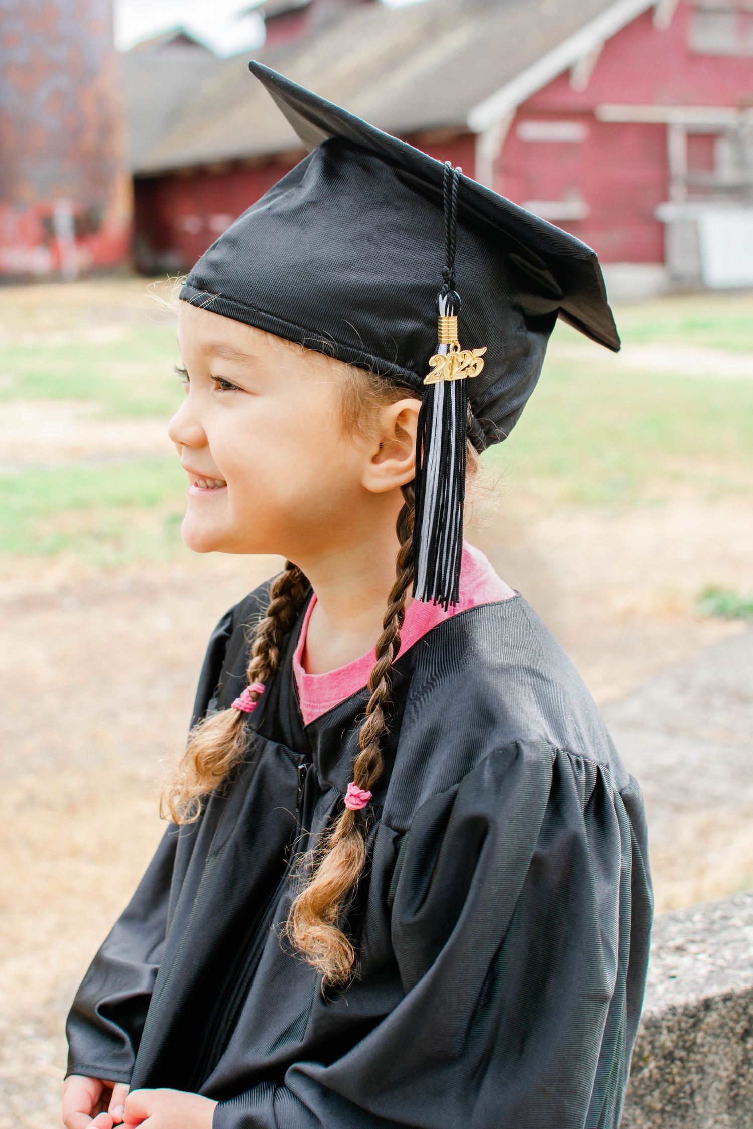 Graduate in black gown and cap with braids smiling in profile against red barn.