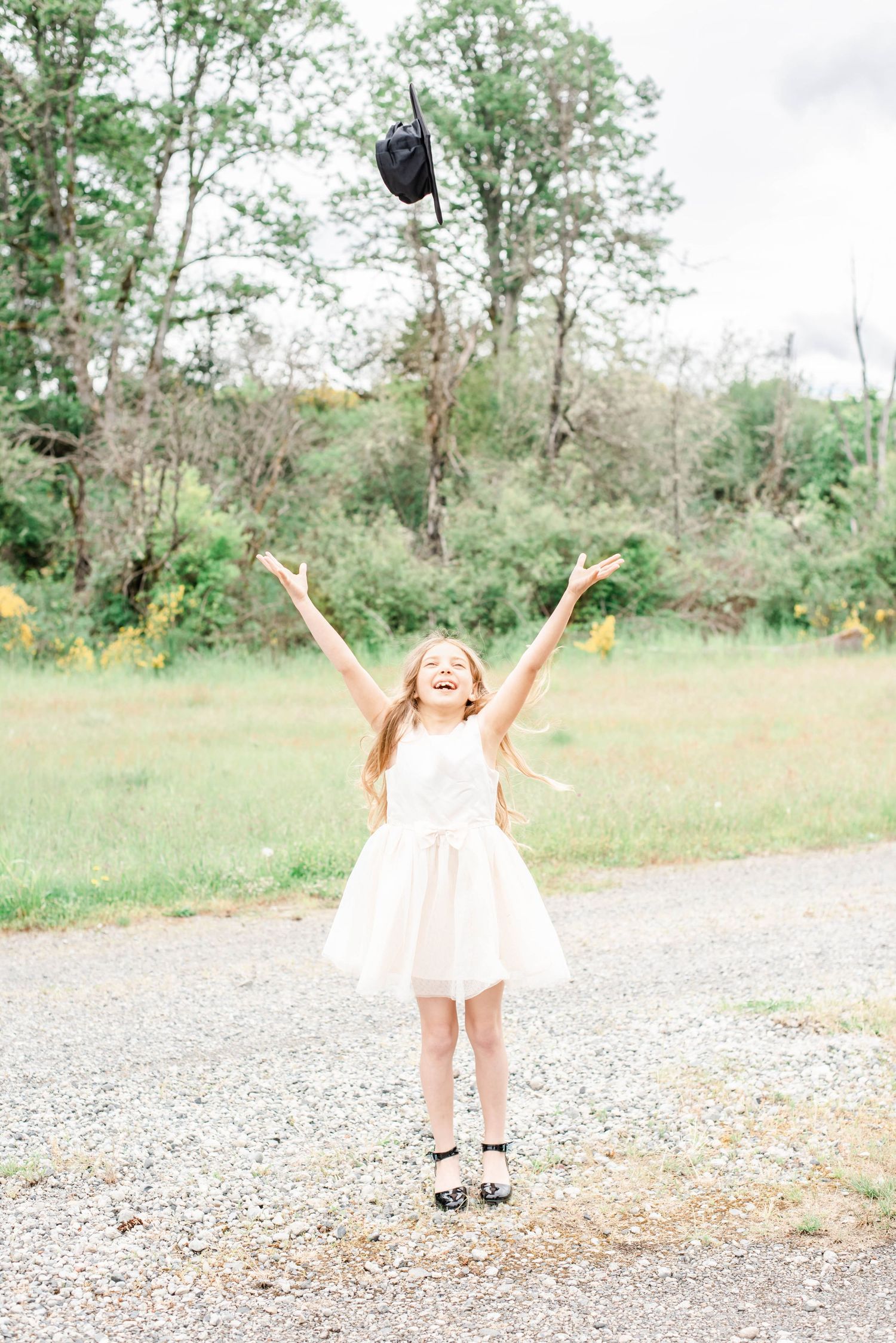 Graduate in white dress joyfully throwing cap in air on gravel path.