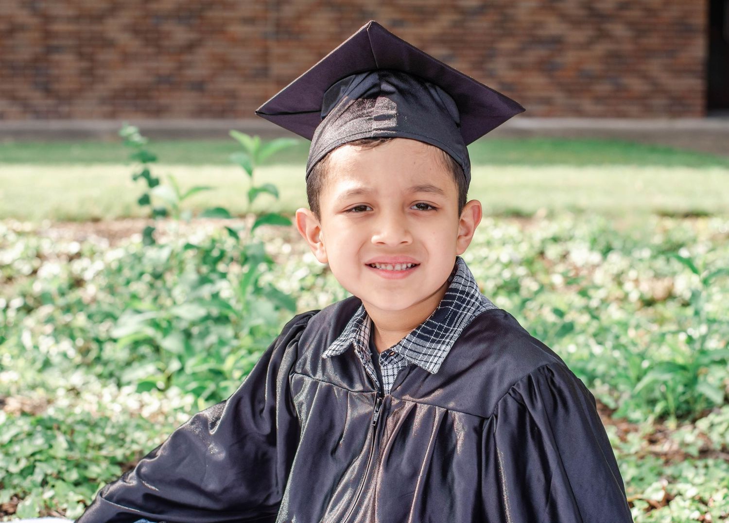 Graduate wearing black cap and gown smiling outdoors with brick wall background.