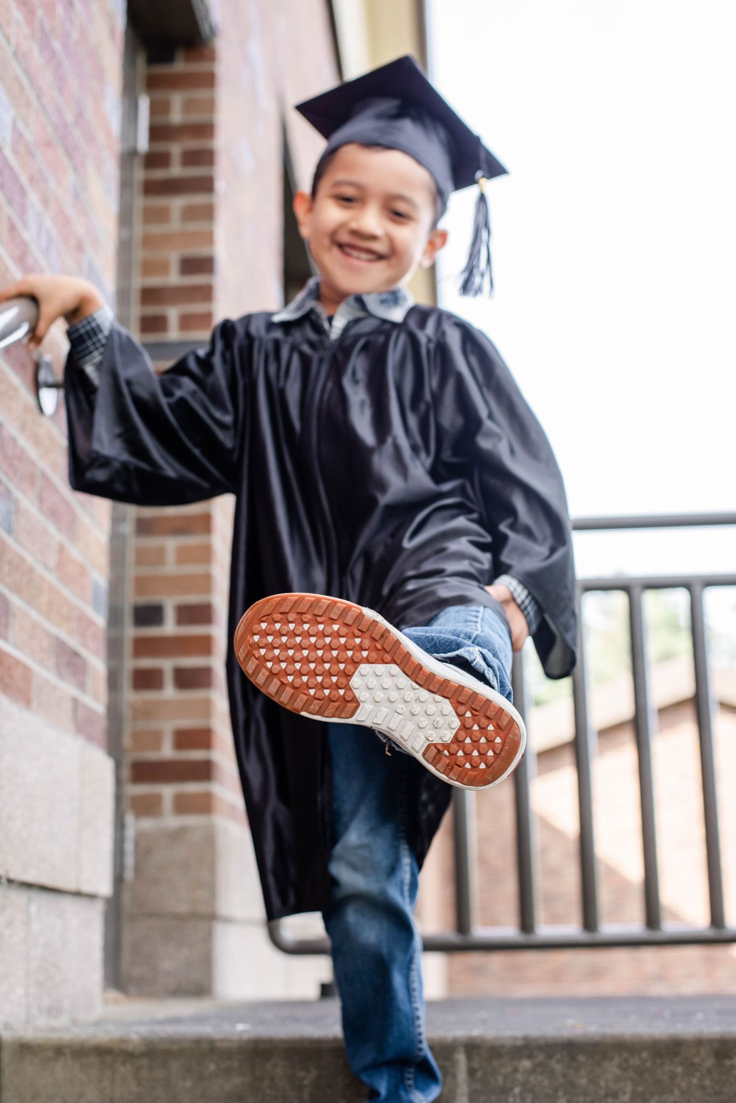 A graduate in black cap and gown shows off playful kicks while posing near brick columns.