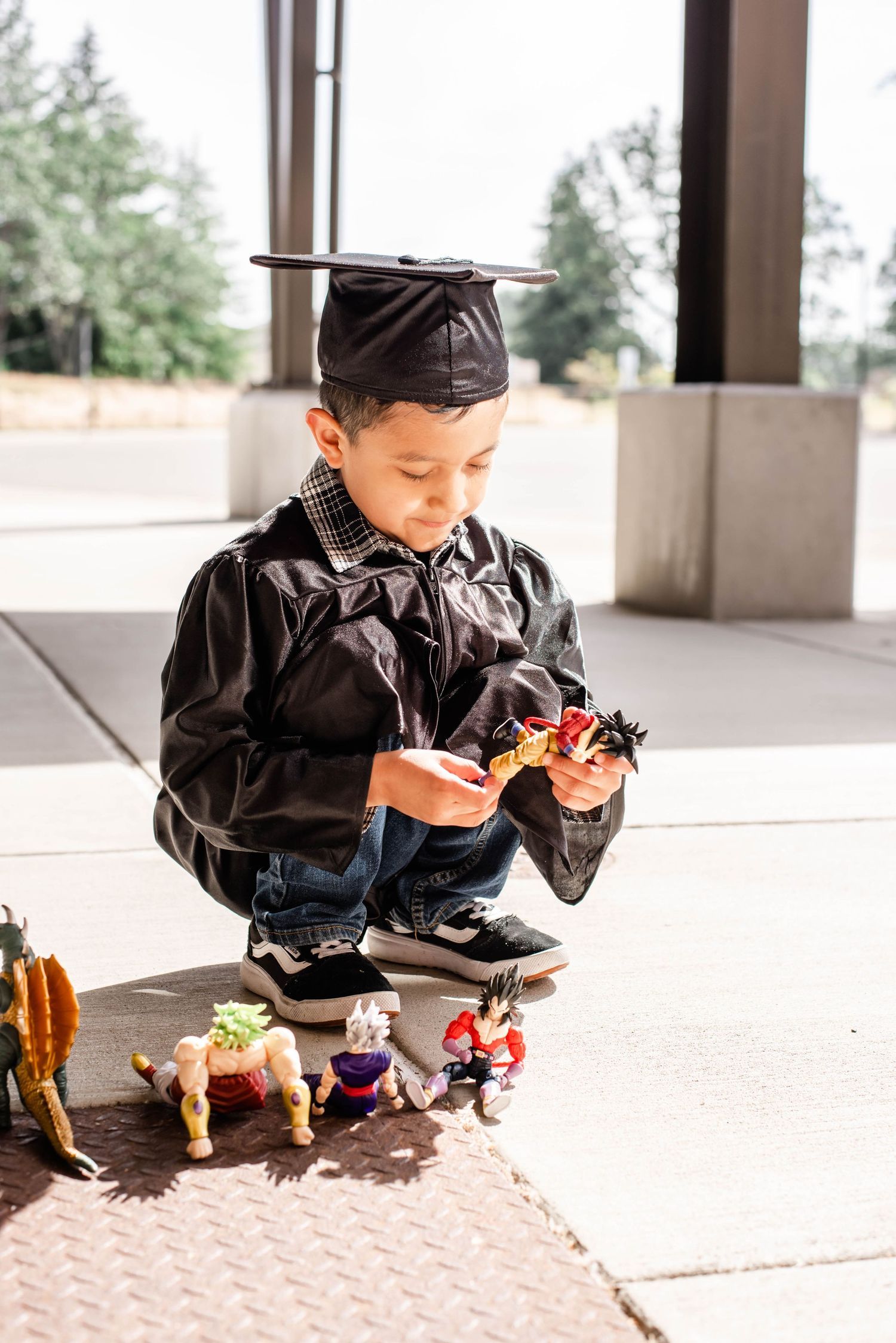 Graduate in black gown and cap playing with colorful toy figures on concrete.