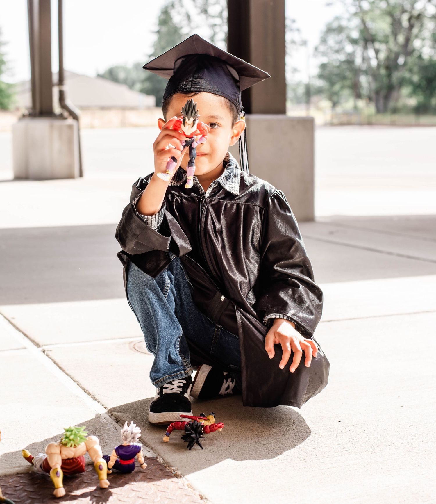 Graduate in black cap and gown playing with toy figures on concrete ground.