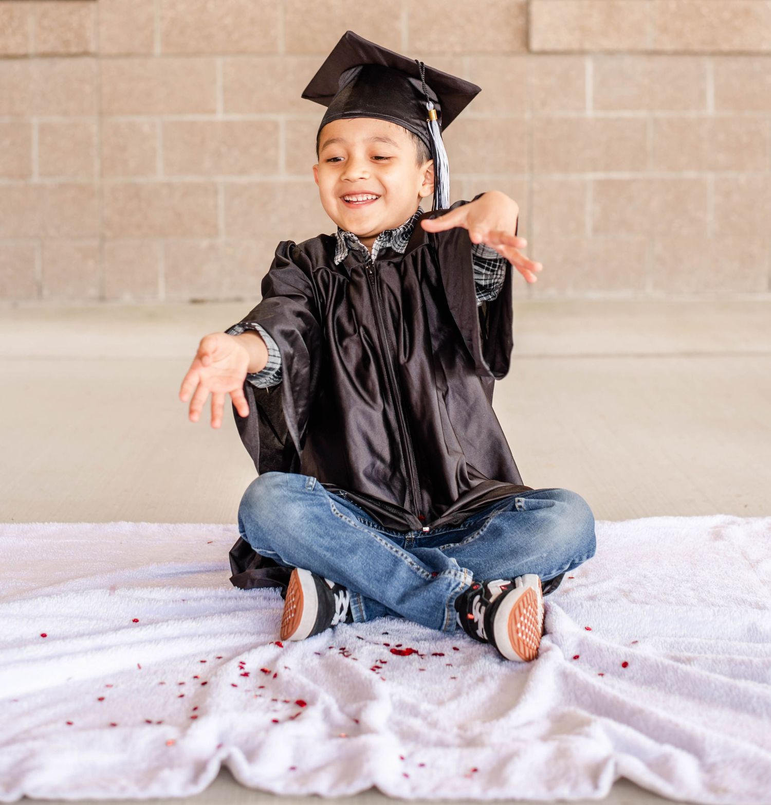 Young graduate in black gown sitting cross-legged on white blanket with confetti.