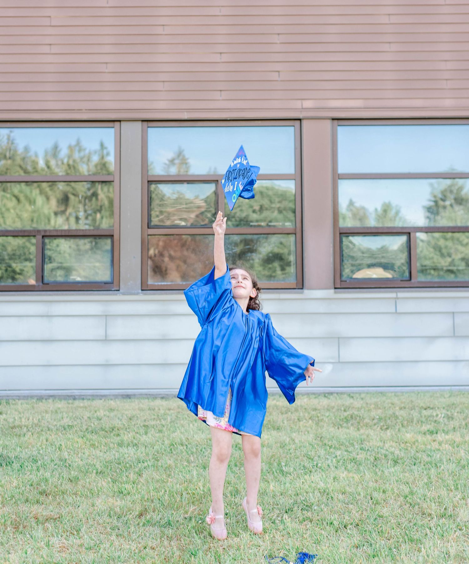 A graduate in blue regalia twirls joyfully on grass outside a school building.