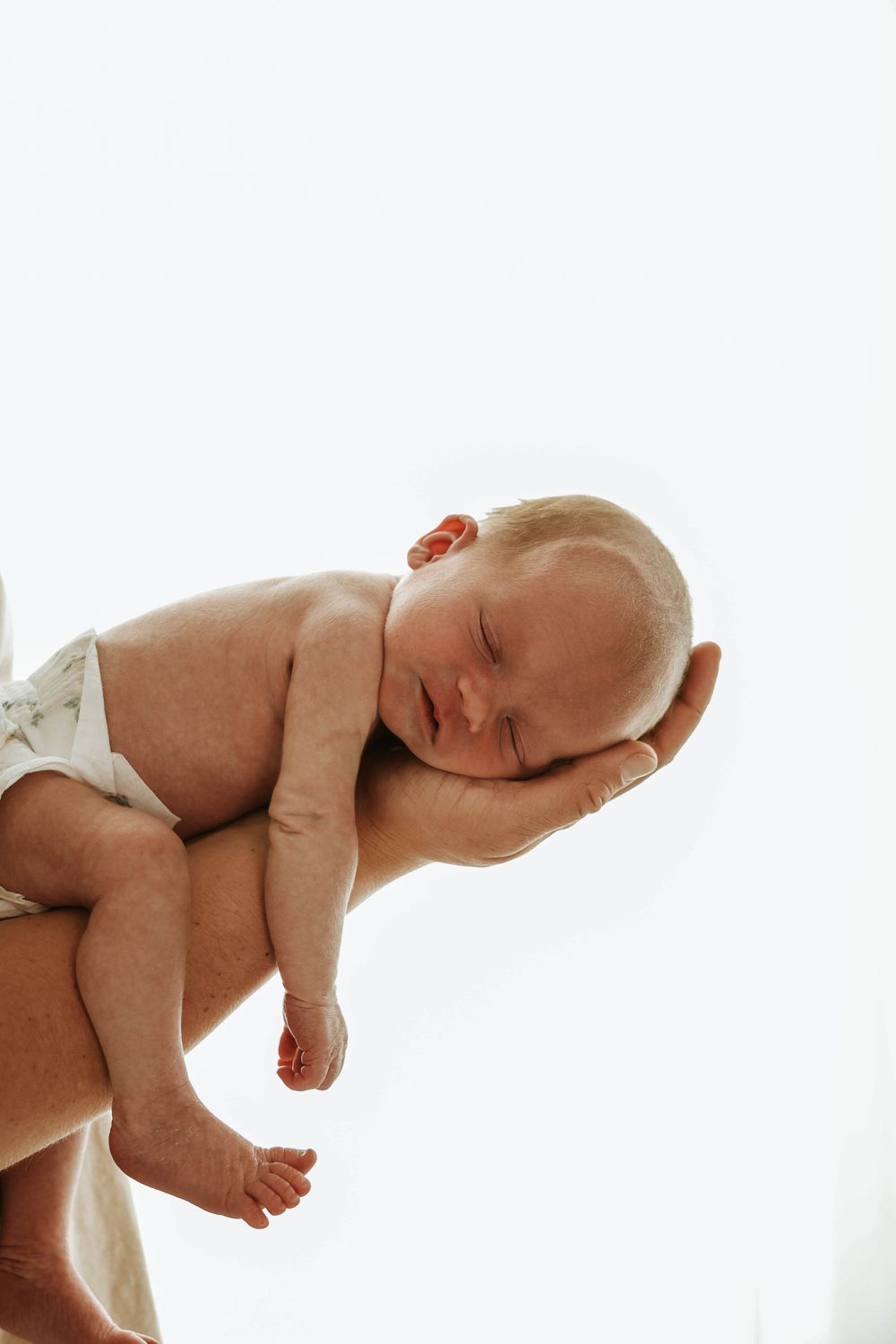 A newborn baby wearing a diaper rests peacefully in an adult's raised hands against a white background.