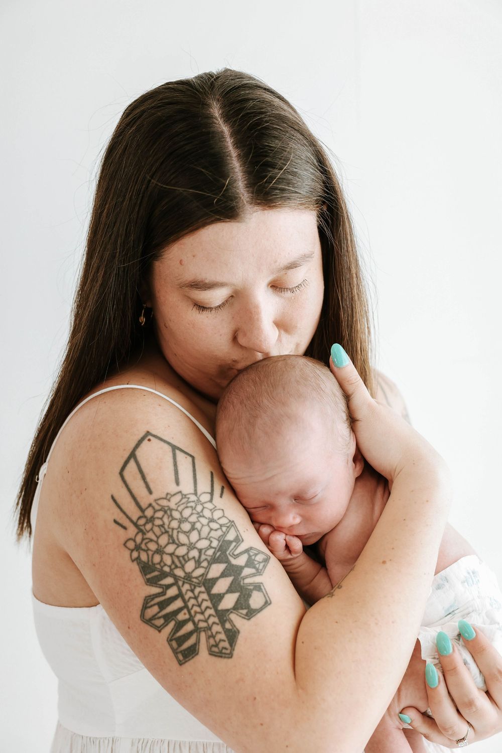 A tender moment shows a person with a floral arm tattoo embracing and kissing a sleeping newborn baby.