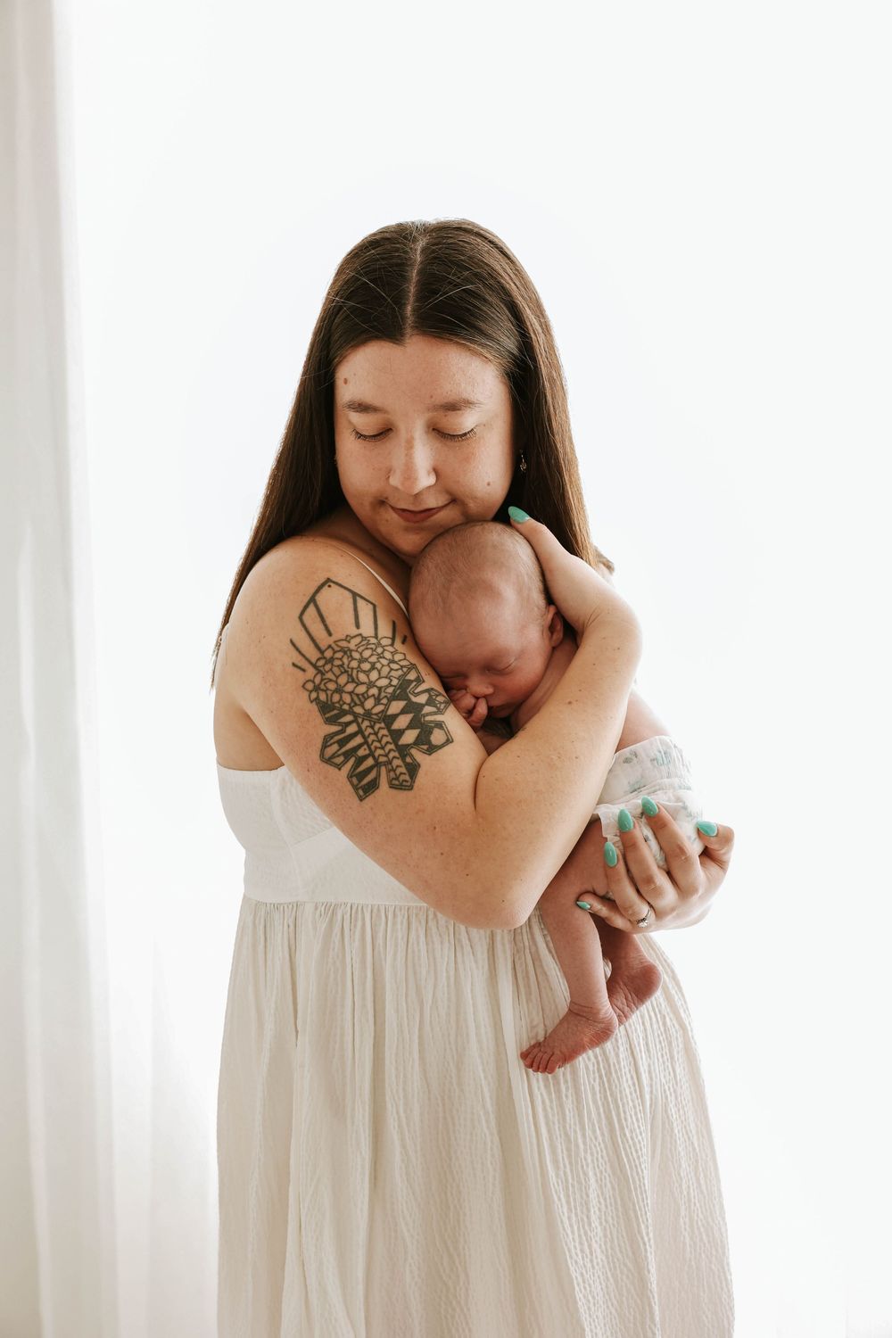 Woman in white dress holding sleeping newborn baby against white curtain background, tattoo visible on her arm.