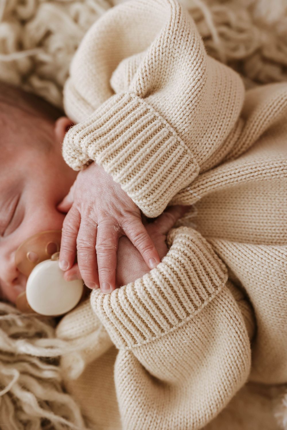 Tiny baby hand reaching out while wearing a cozy cream colored knitted sweater against neutral textured background.