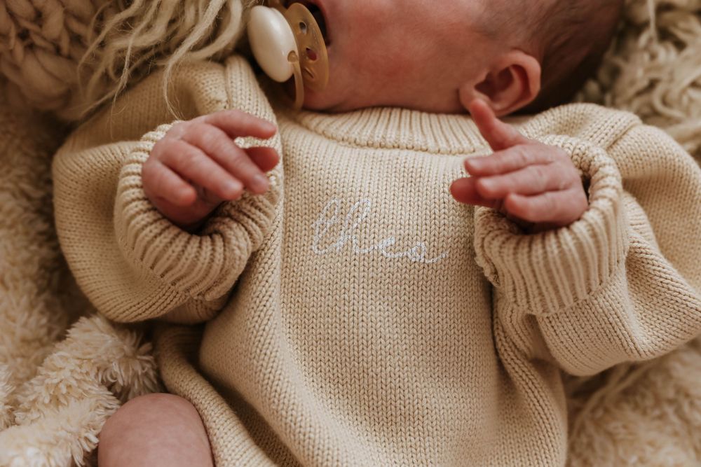 Close-up of tiny baby hands grasping a beige knit sweater with delicate details.