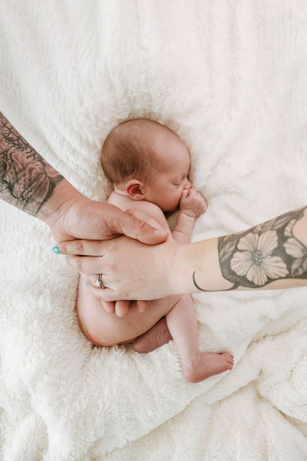 Gentle tattooed hands cradle a sleeping newborn baby on soft white blankets.