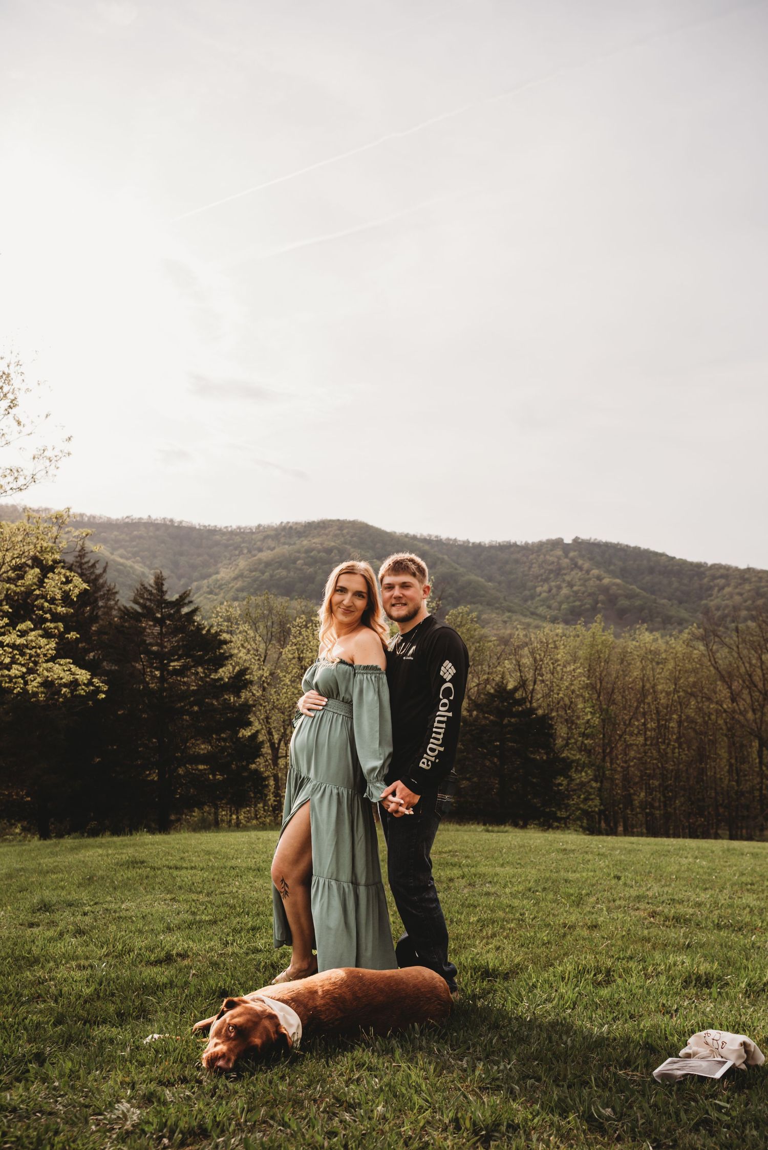 Couple stands in mountain meadow with their resting dog at their feet.
