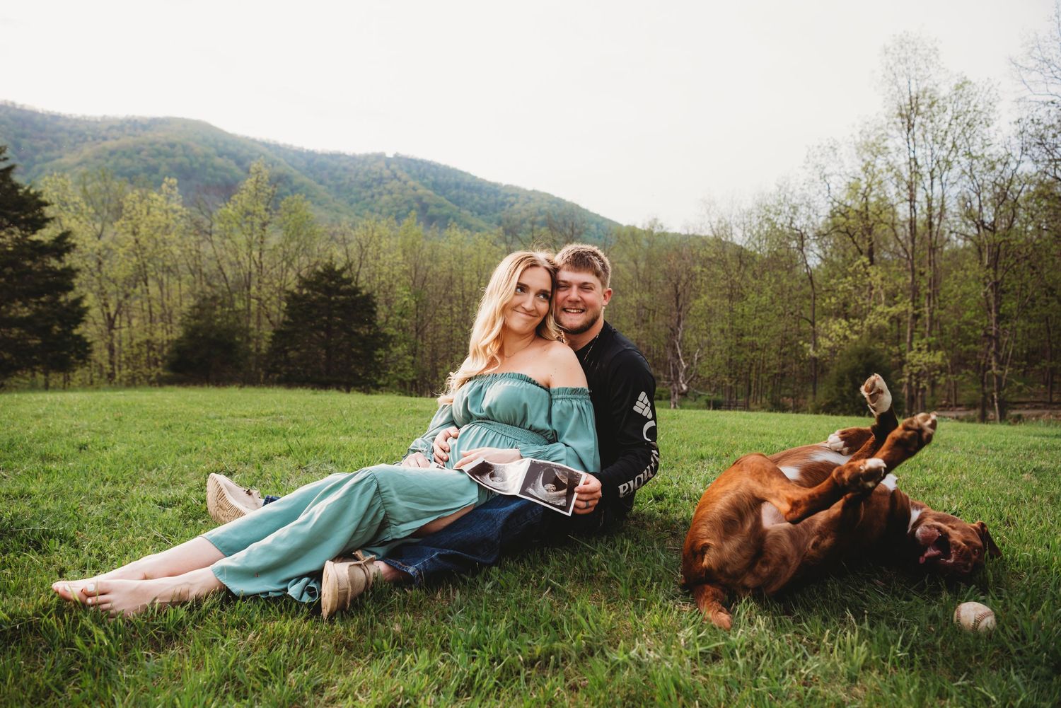 Couple relaxes in mountain meadow with playful dog rolling nearby.